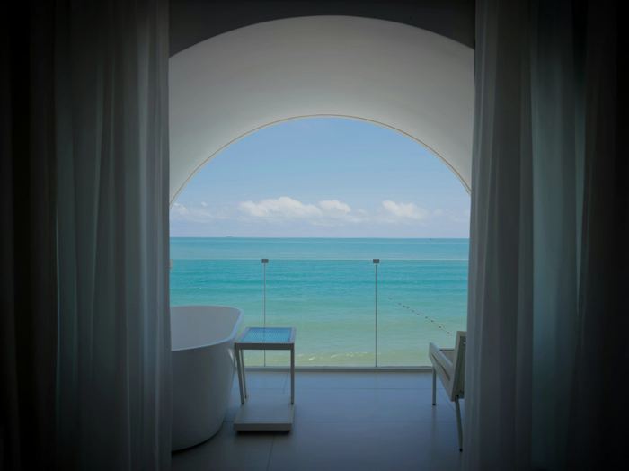 A white bath at a tall window, the turquoise sea framed by the curtains.