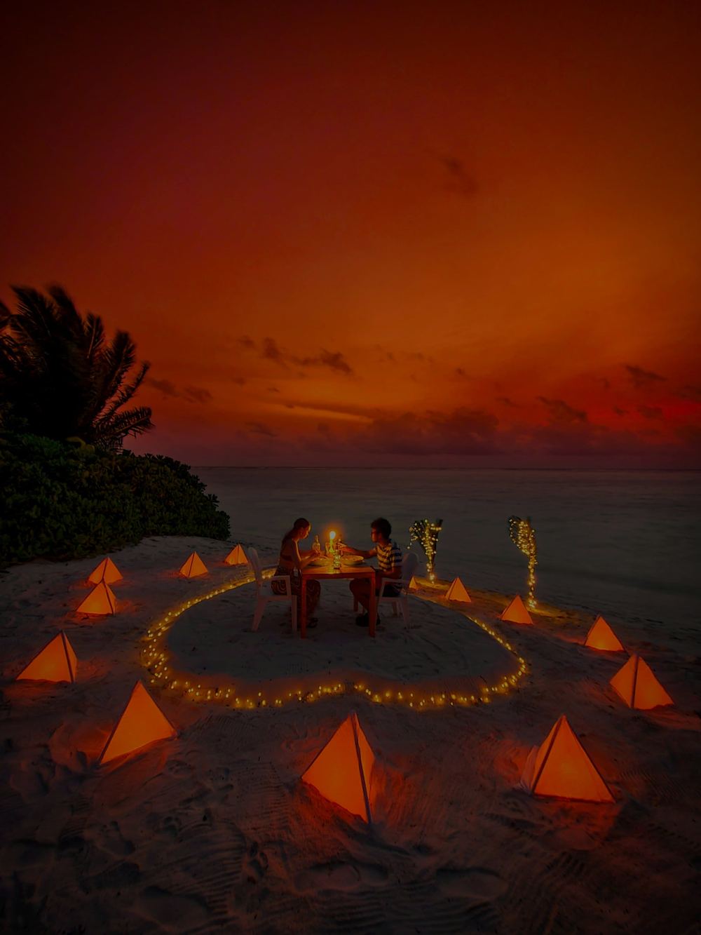 A couple at a candlelit table on the beach at sunset, lanterns set in a ring around them.