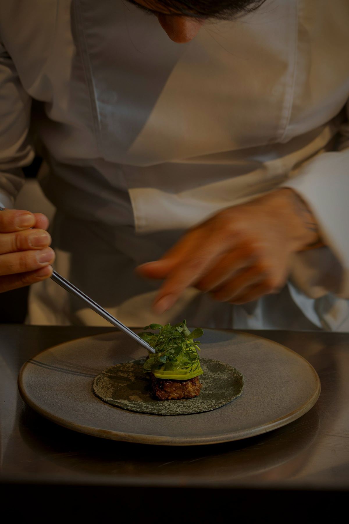 A chef in whites plating a small dish with tweezers.