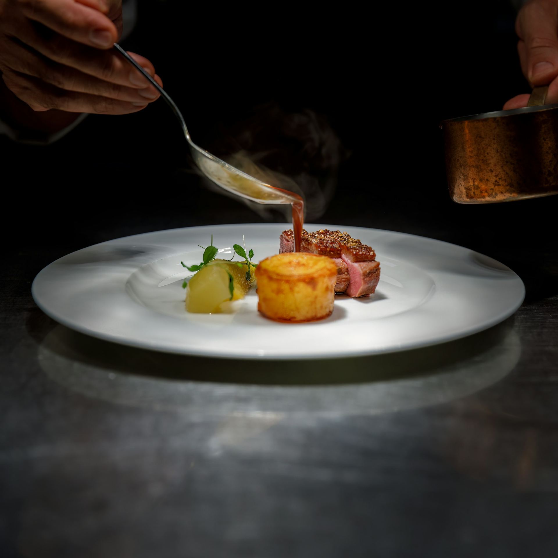 A chef pours sauce over a plated dish on a white plate, in low light.