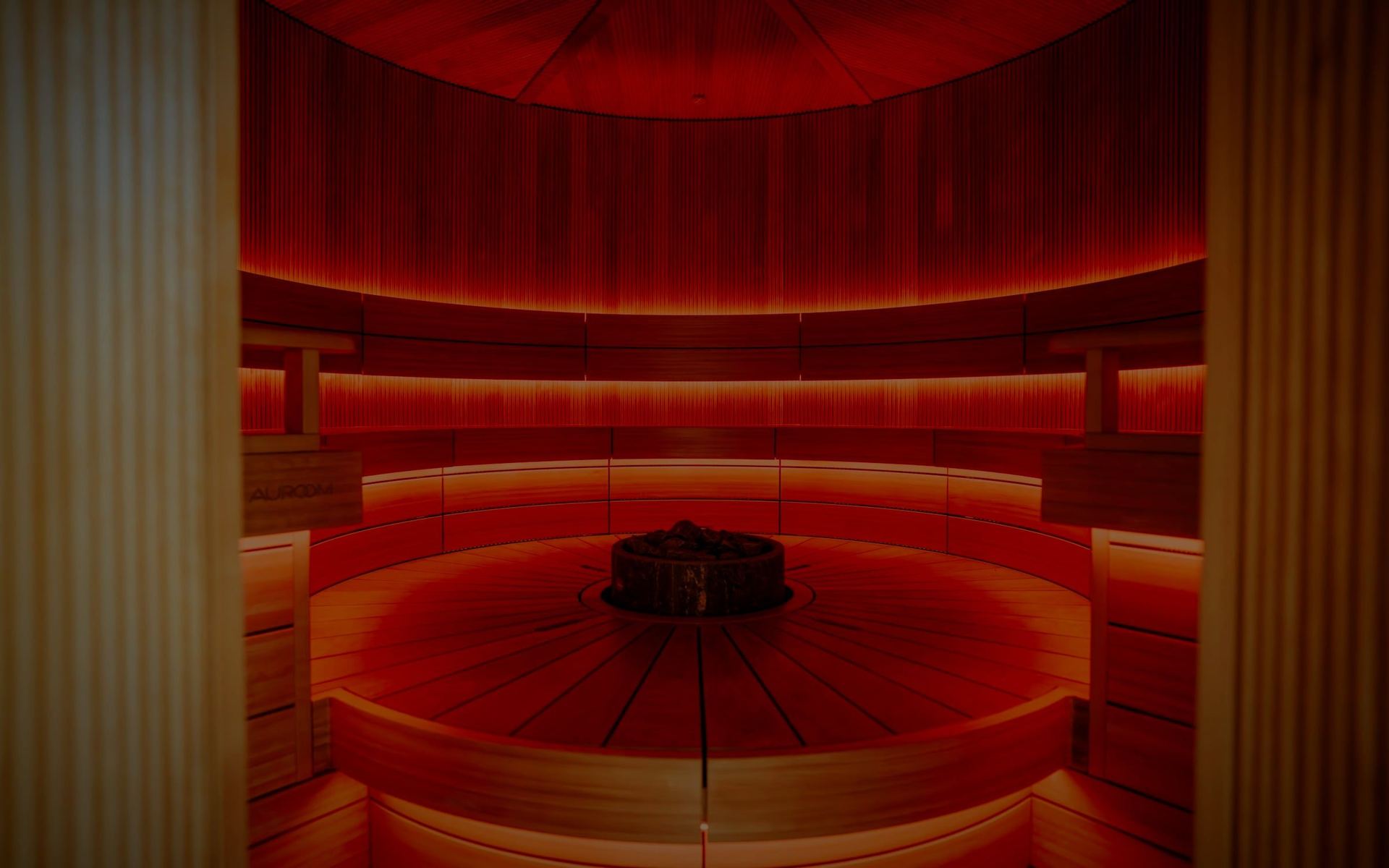 A round heat room lined in timber and lit a deep red, a basket of stones at its centre.