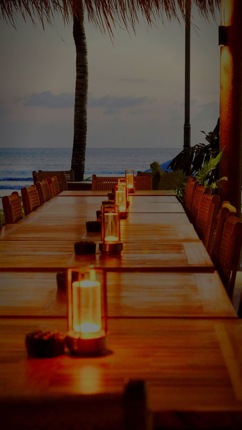 A long wooden table set with candles in glass, palms and the sea at dusk.