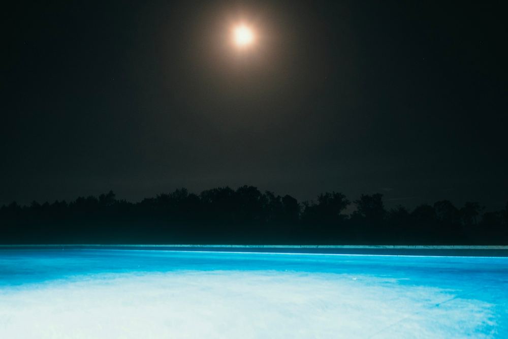 An infinity pool glowing blue under a full moon, the tree line dark beyond.