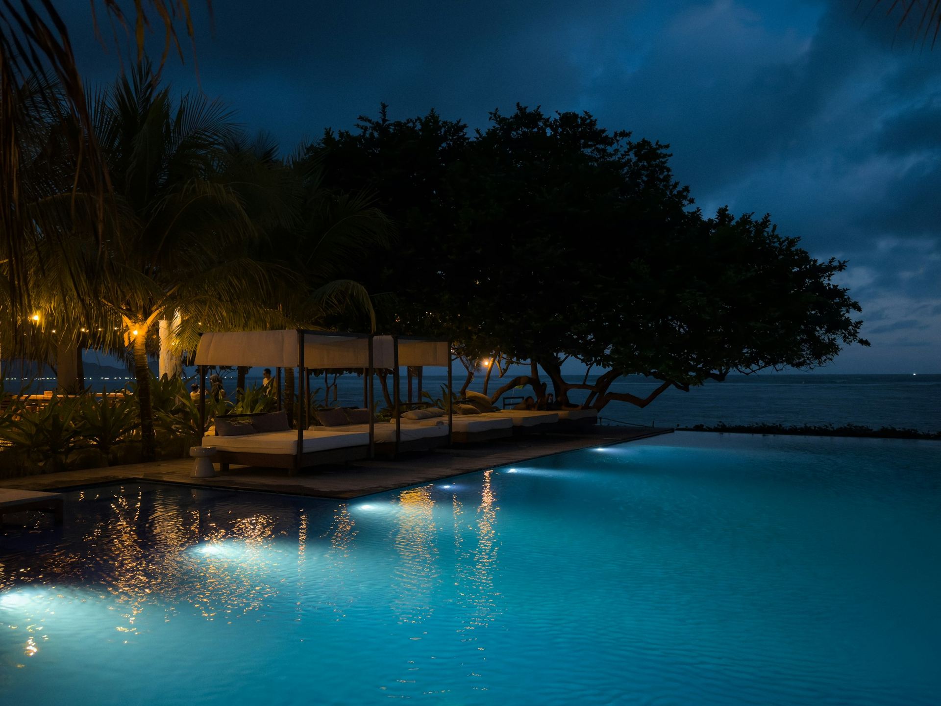 An infinity pool lit blue at dusk, a cabana and great trees at its edge and the sea beyond.