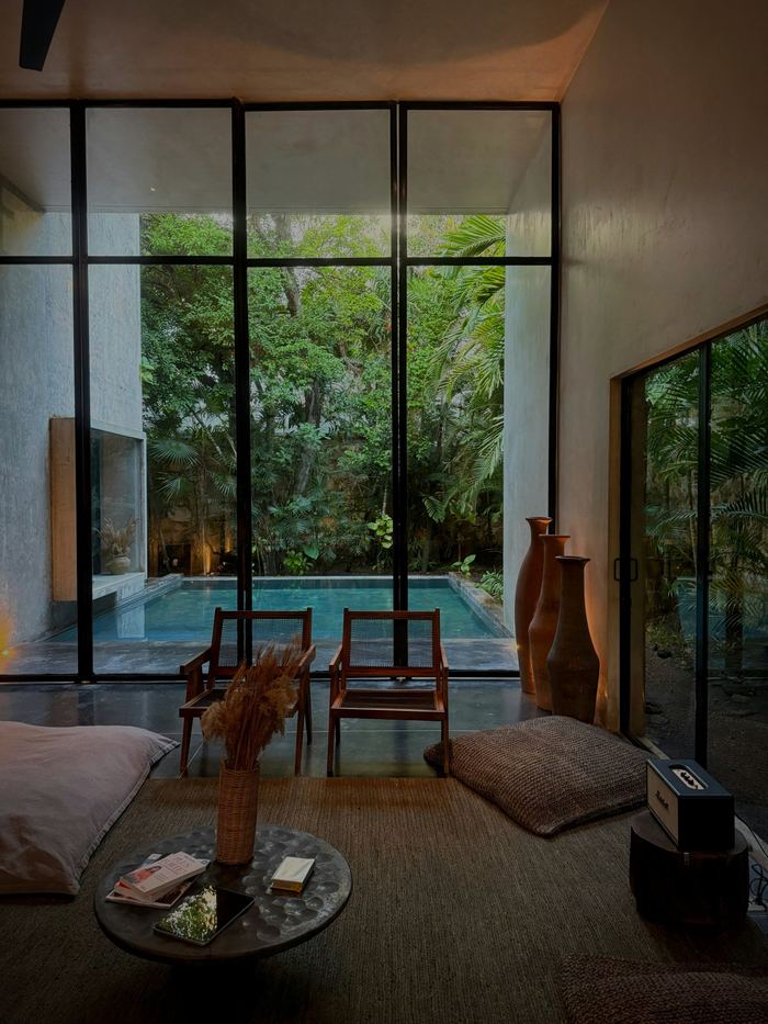A living room with a long low sofa, glass walls open to a pool and dense green garden.