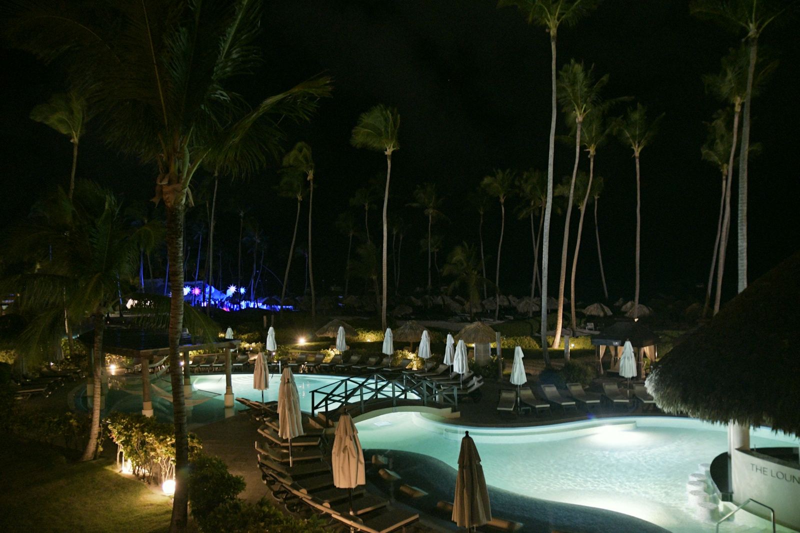 Pools lit blue among tall palms at night, parasols folded and lamps along the paths.
