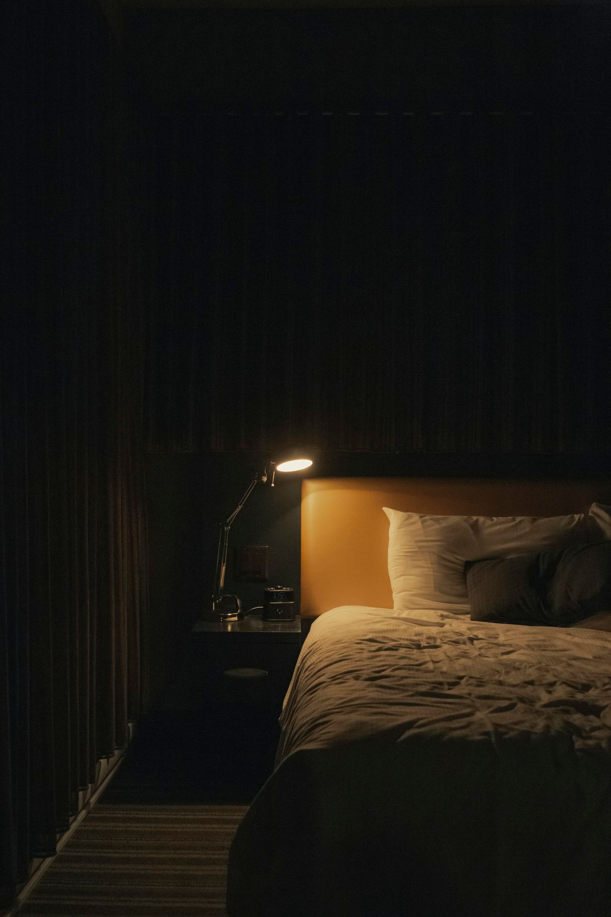 A bed under a single reading lamp, dark curtains drawn and the room in low gold light.