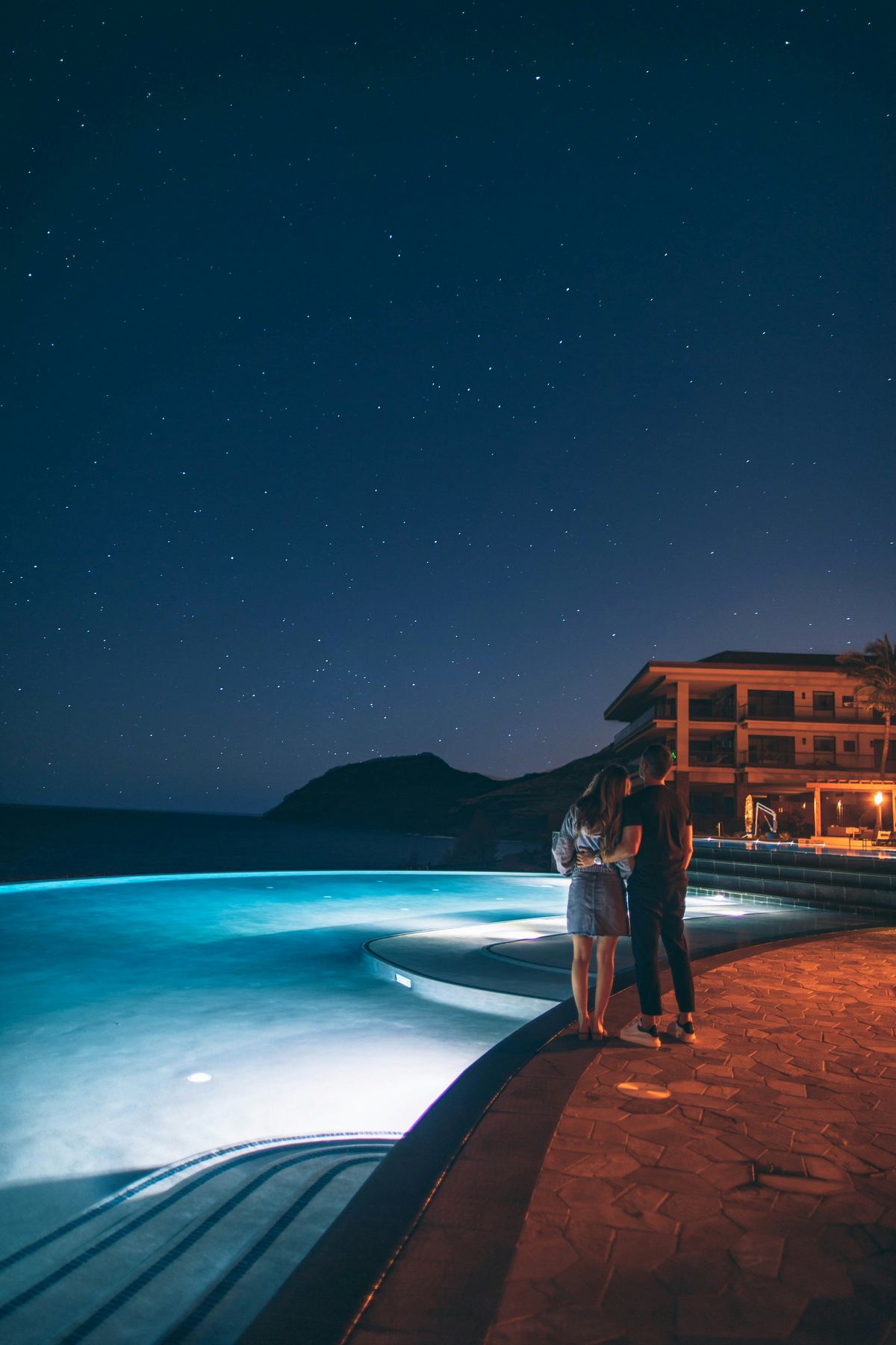 Two guests at the edge of a lit infinity pool under the stars, the house glowing behind them and the dark headland beyond.