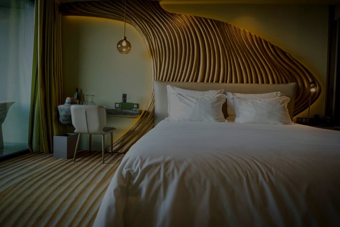 A white bed beneath a sculpted timber headboard, a desk and a window onto green.