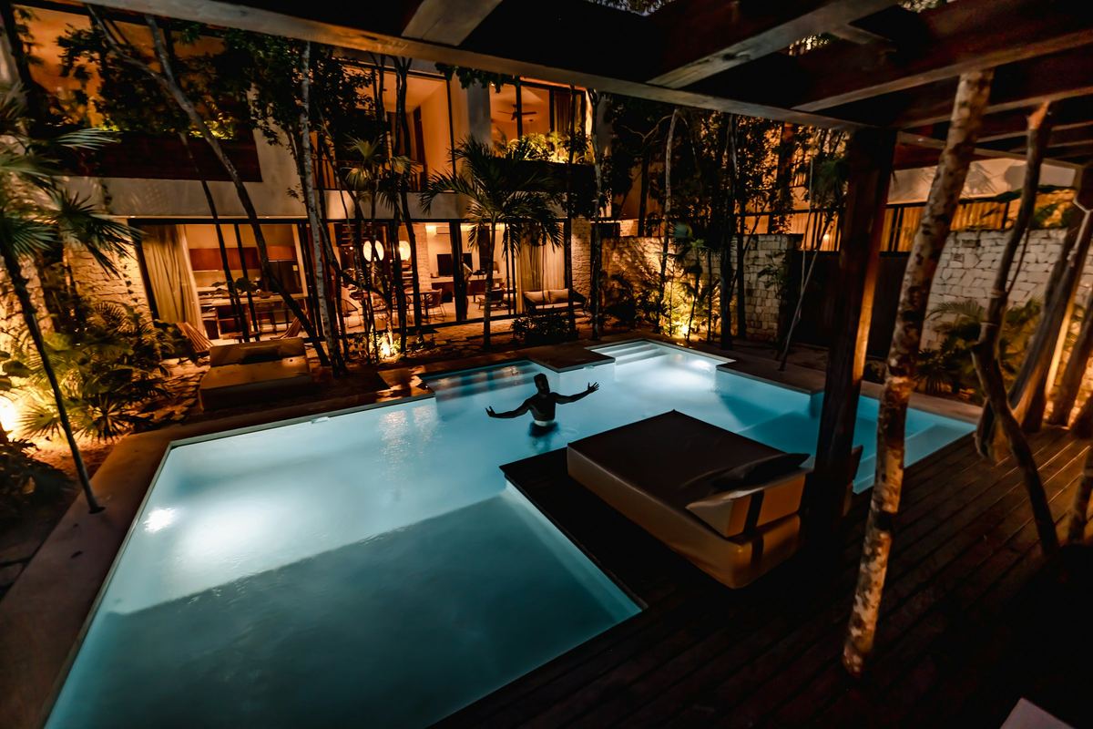 A villa's private pool lit at night under timber eaves and palms, a guest resting at the water's edge.