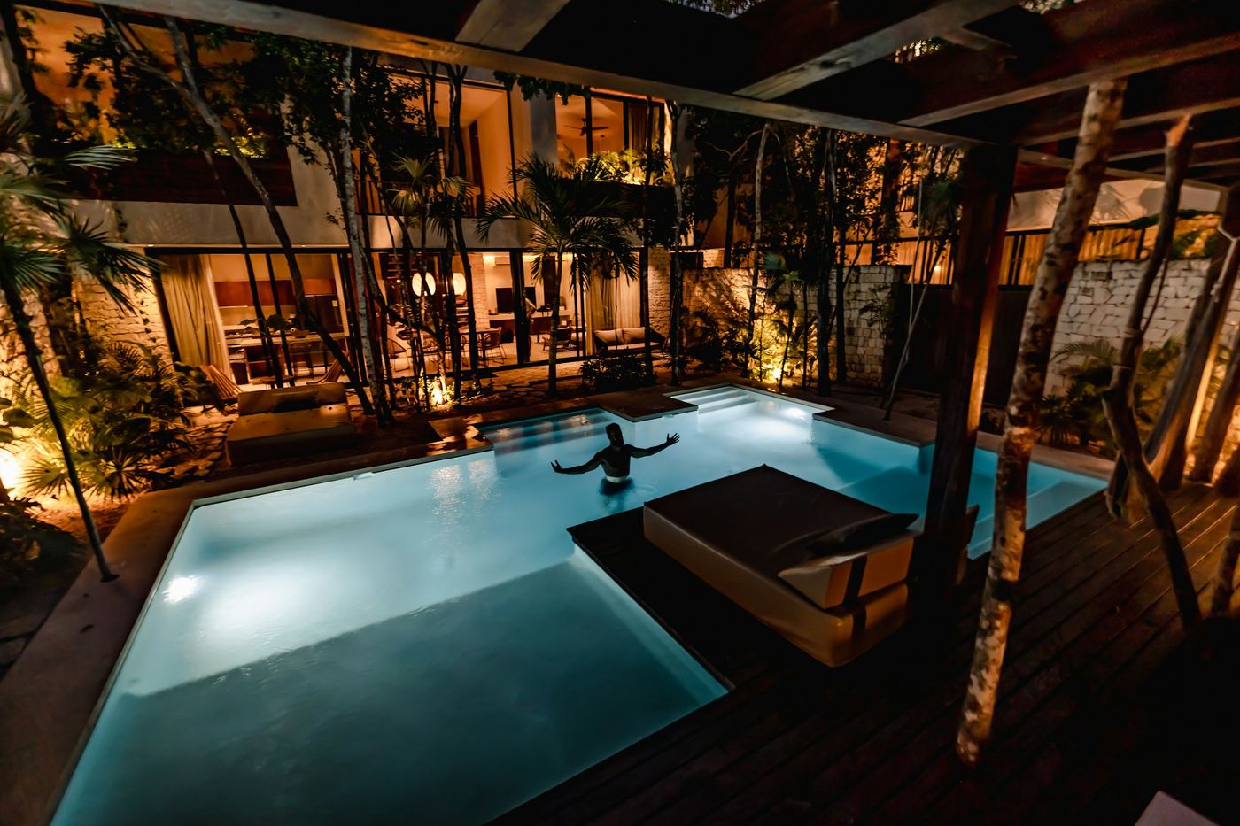 A villa's private pool lit at night under timber eaves and palms, a guest resting at the water's edge.