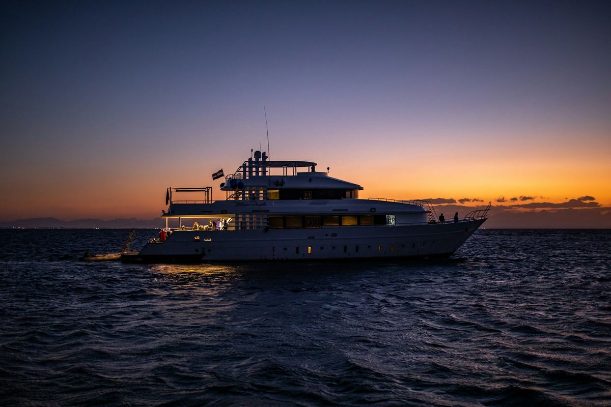 A white motor yacht crossing a dark sea at dusk, the last orange light on the horizon.