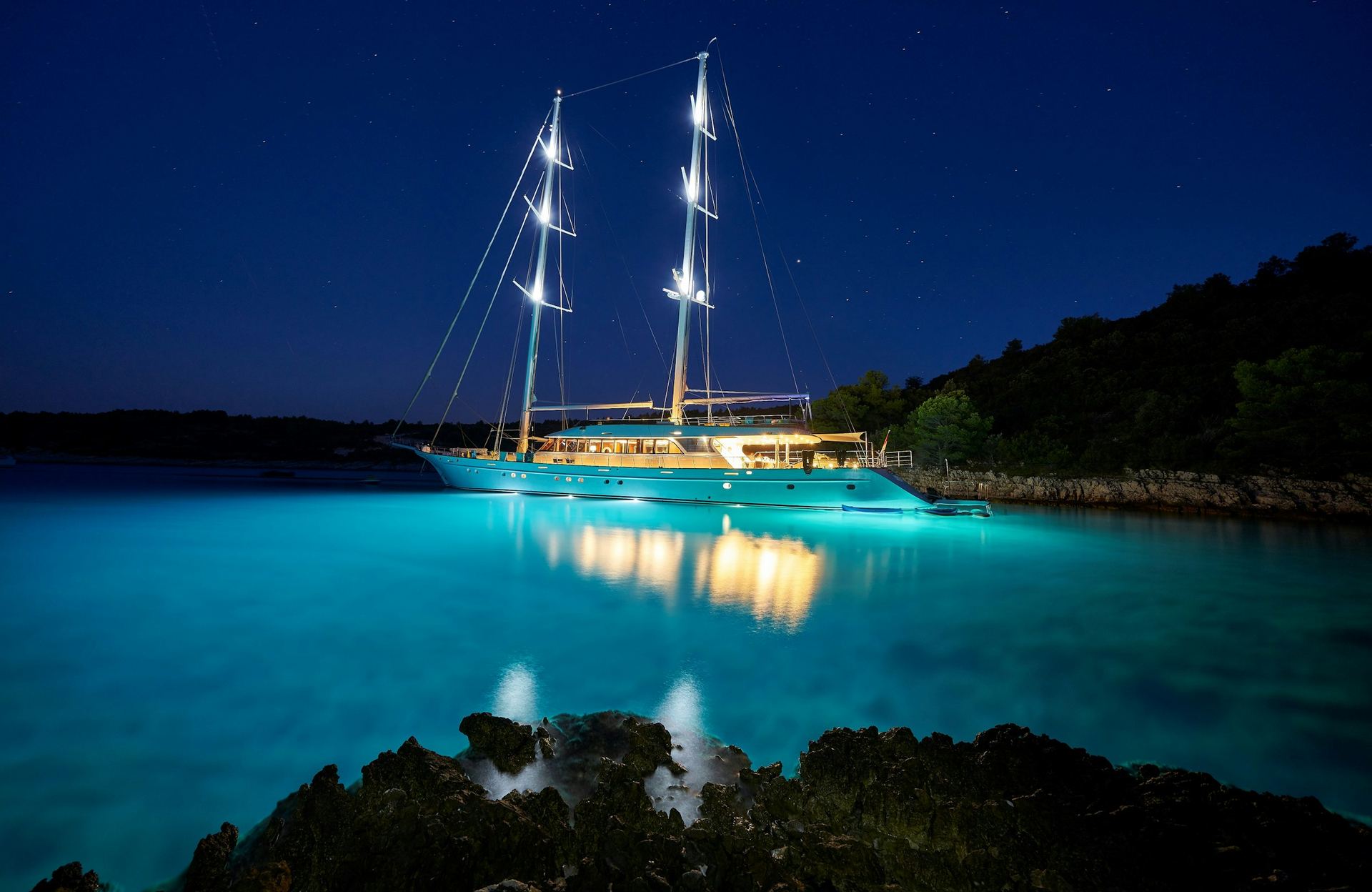 A two masted yacht lit white at anchor in a glowing turquoise cove at night.