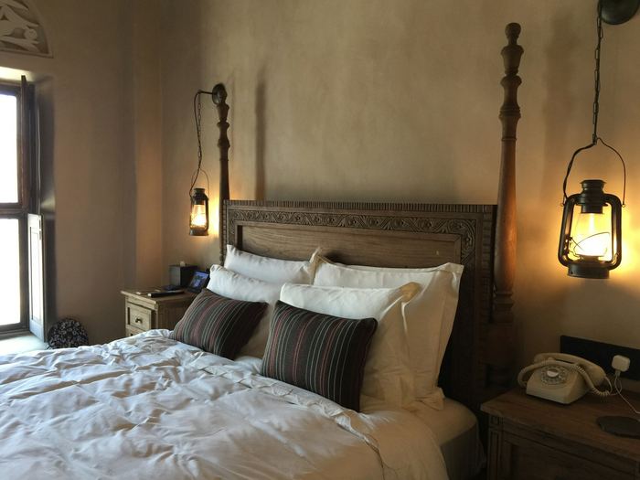 A bed with dark pillows against a carved timber headboard, a hurricane lamp lit beside it.