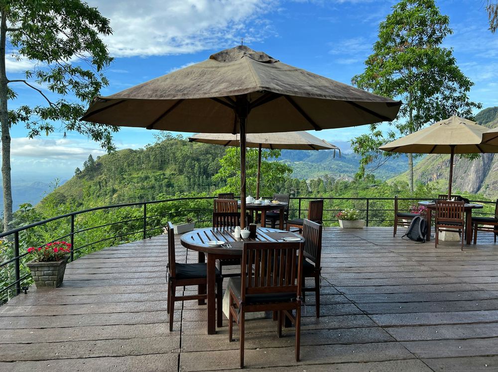 A dining table under a parasol on a timber deck above forested hills.