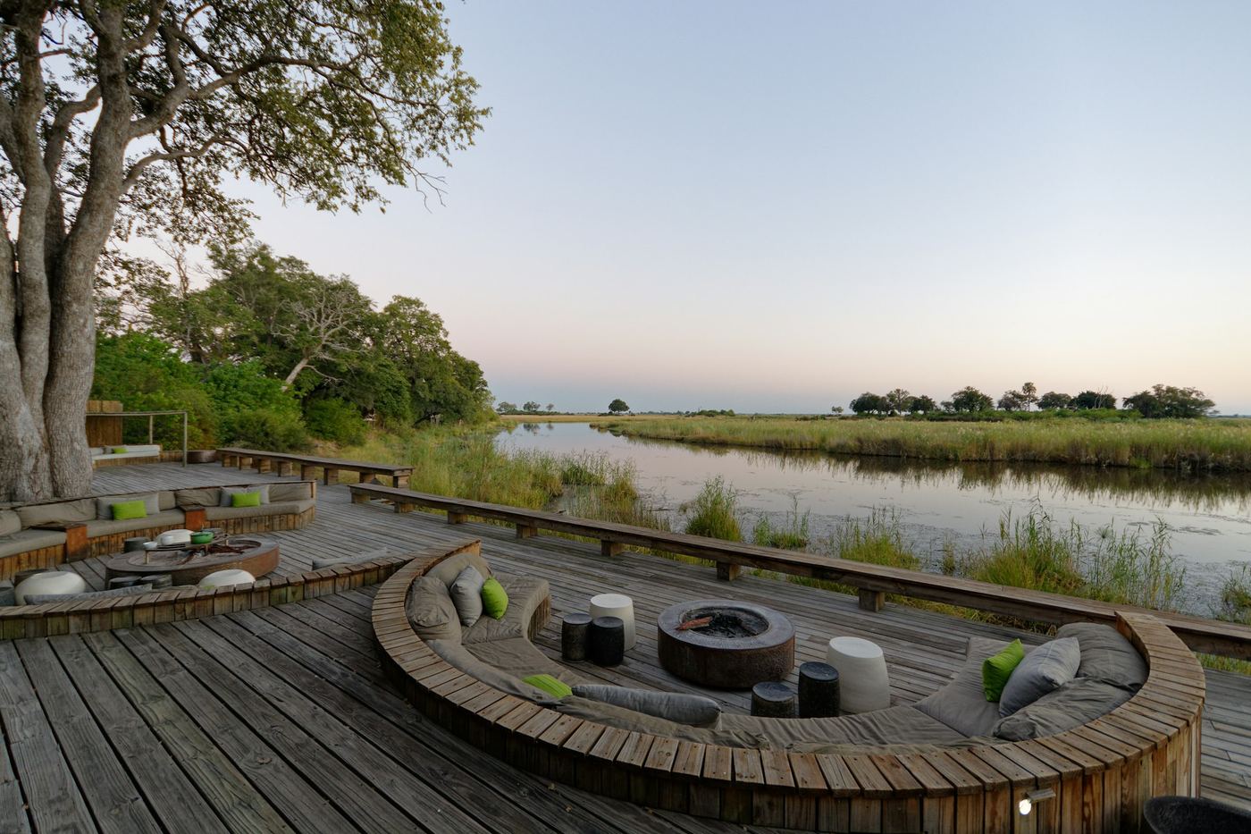 A timber deck with a sunken fire pit beside a still river, trees along the far bank.
