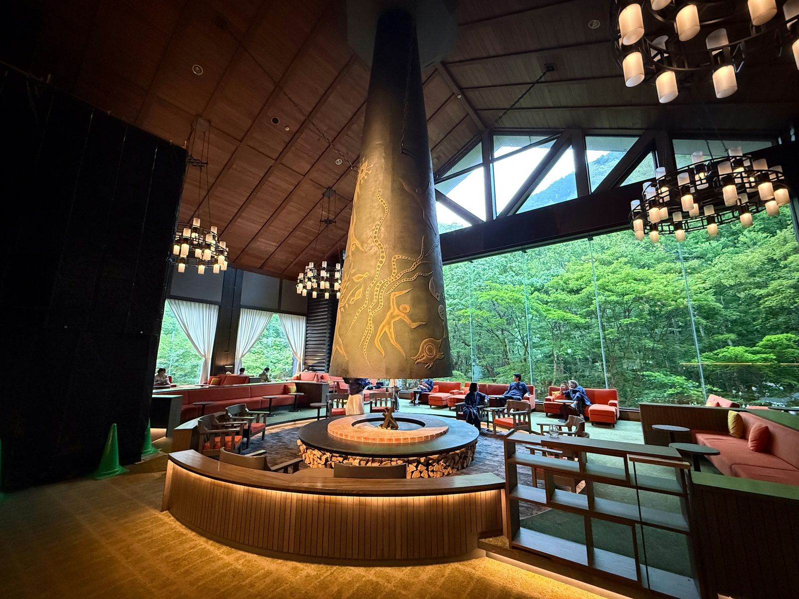 A lodge lounge with a great conical fireplace, low sofas and a wall of glass onto the forest.