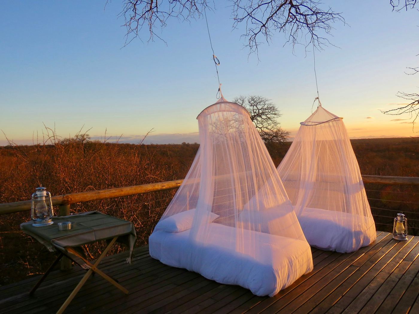 Two beds under white mosquito nets on a timber deck, the plains behind them at sunset.