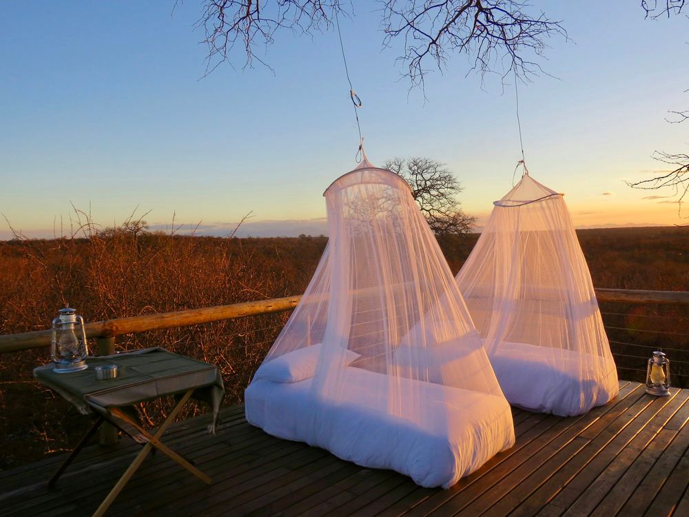 Two beds under white mosquito nets on a timber deck, the plains behind them at sunset.