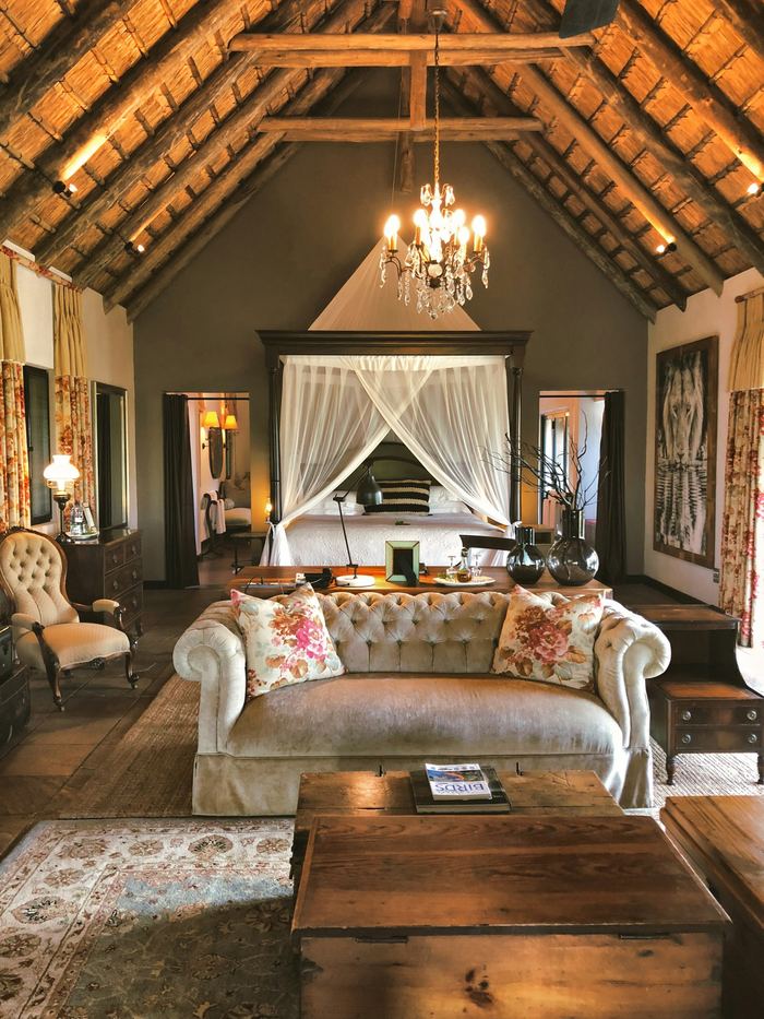 A suite with a tufted sofa, a four poster bed under white drapes and dark timber walls.