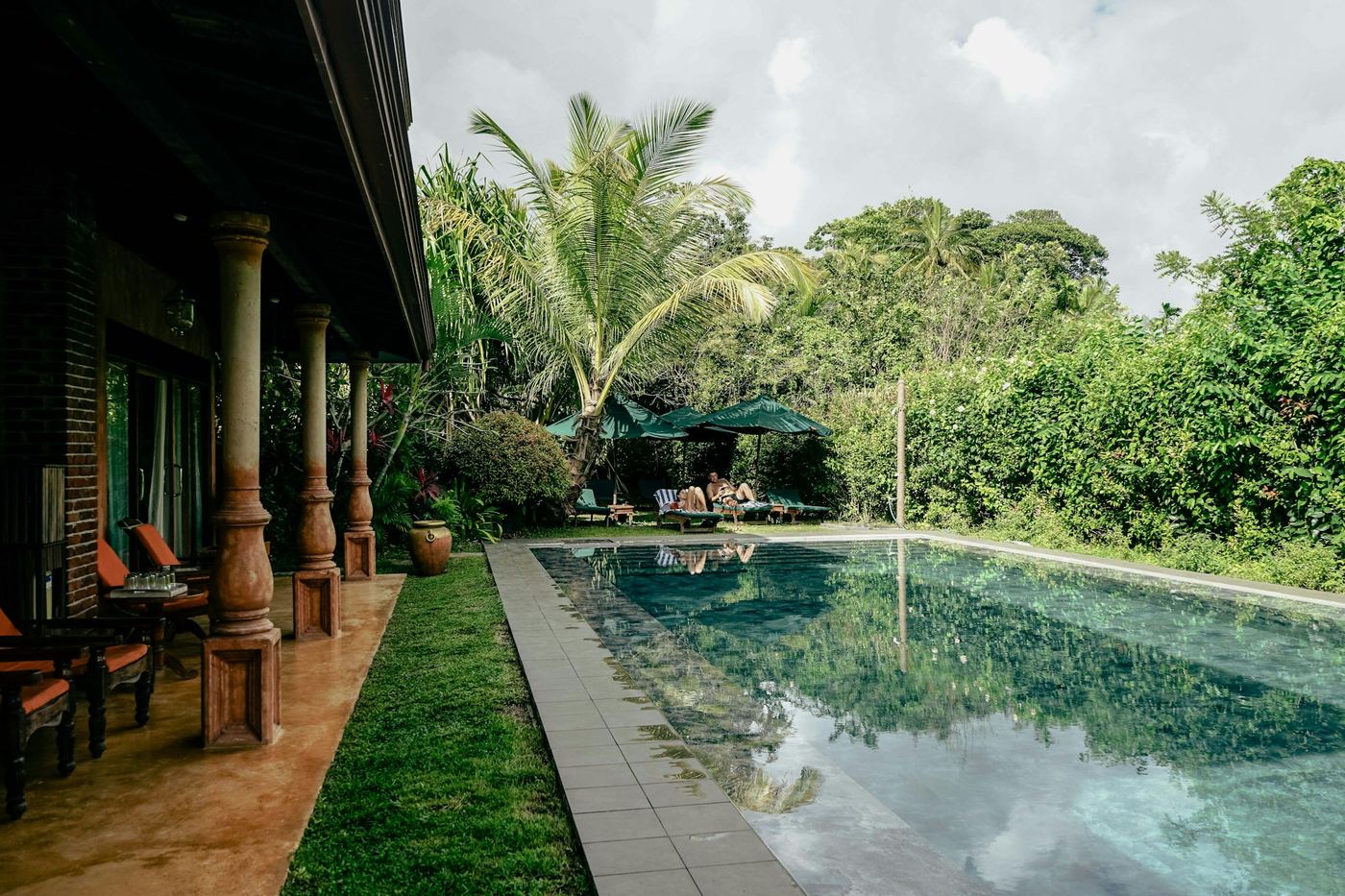 A long pool beside a veranda of carved pillars, a lawn and dense green garden beyond.