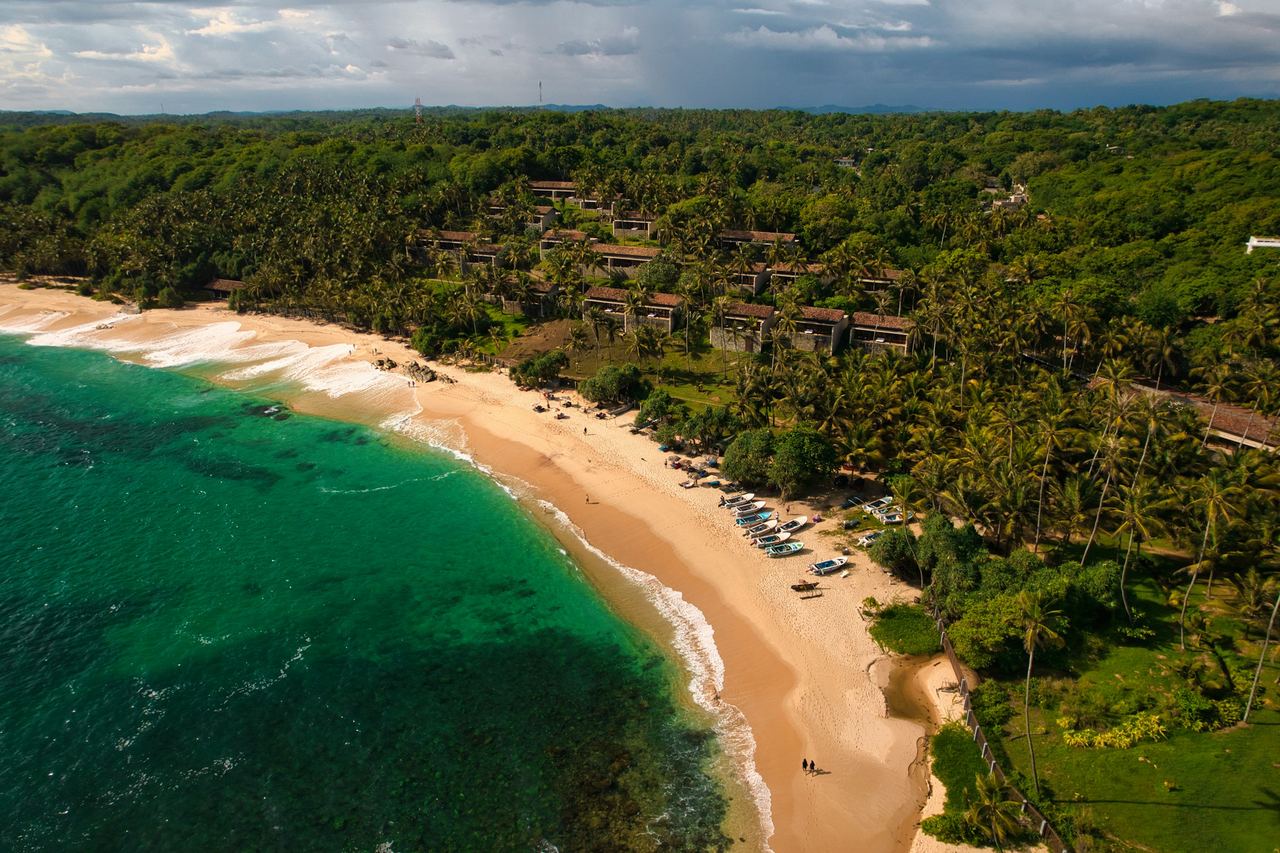 A resort among the palms above a curved beach and a turquoise bay on the south coast, photographed from the air.
