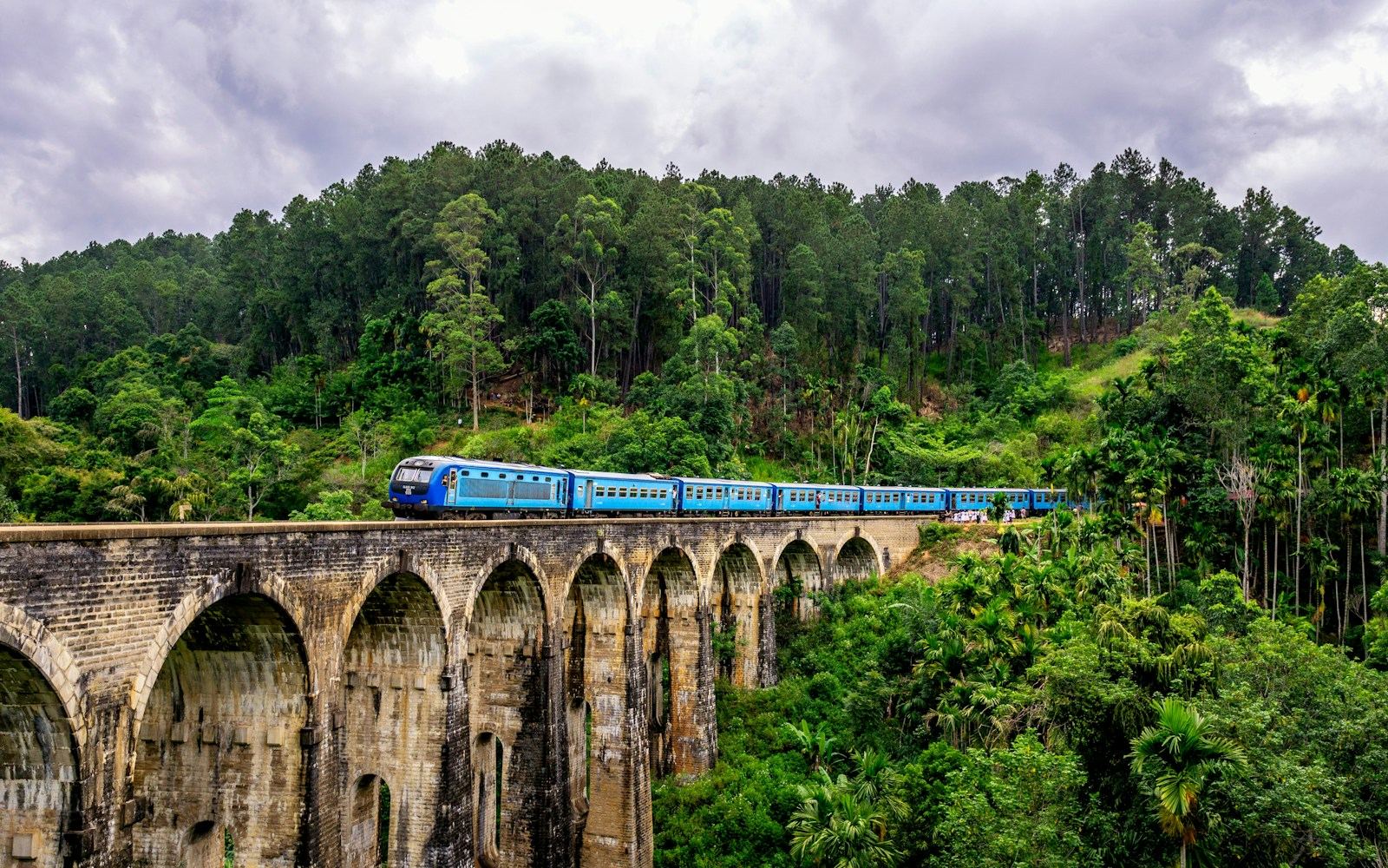 A blue train crossing the stone arches of the Nine Arch Bridge through the forest at Ella.