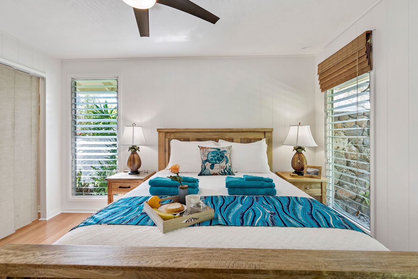 A white bedroom with louvred windows, a ceiling fan and a teal throw on the bed.