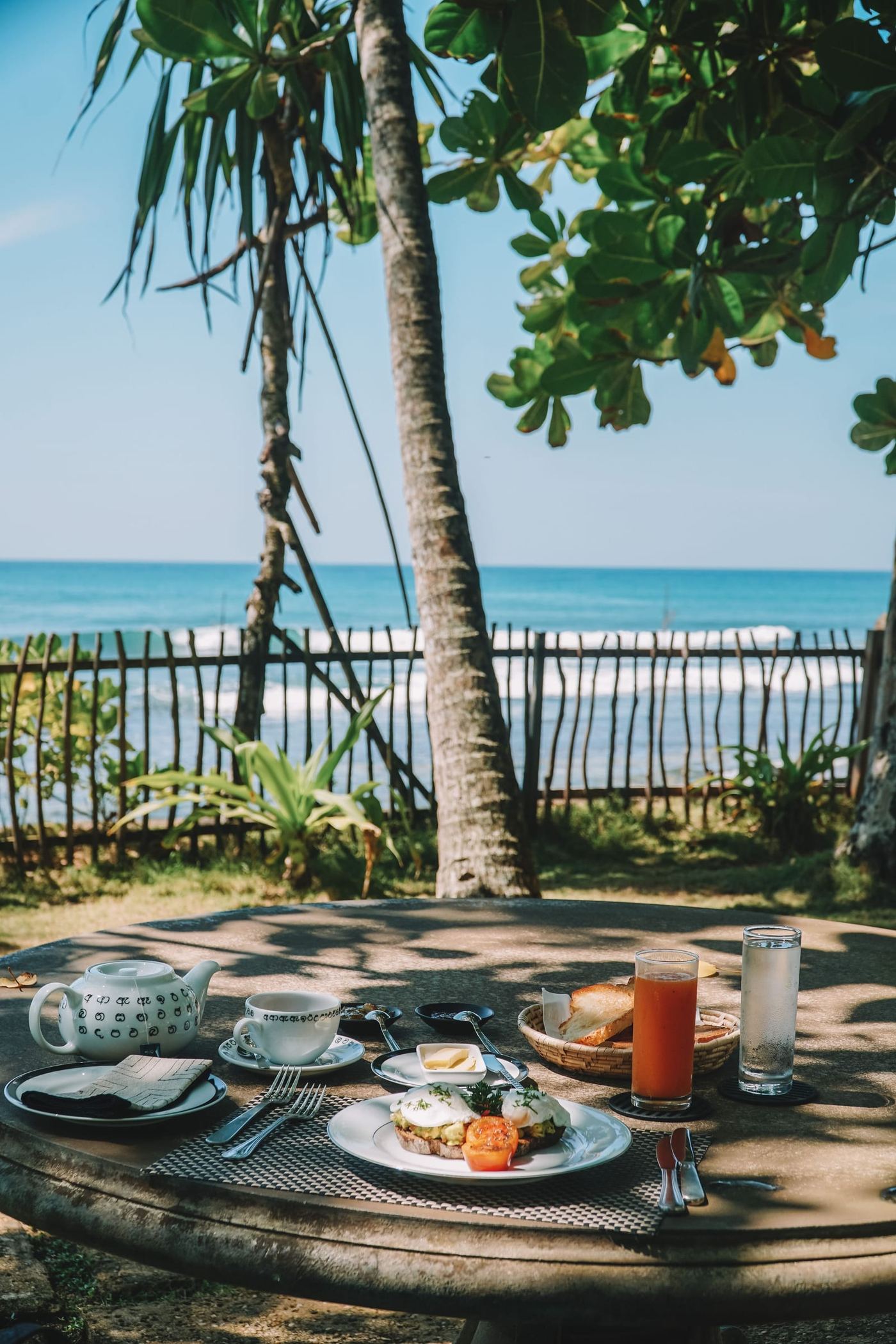 Breakfast laid on a table by a low fence, palms and the sea behind.
