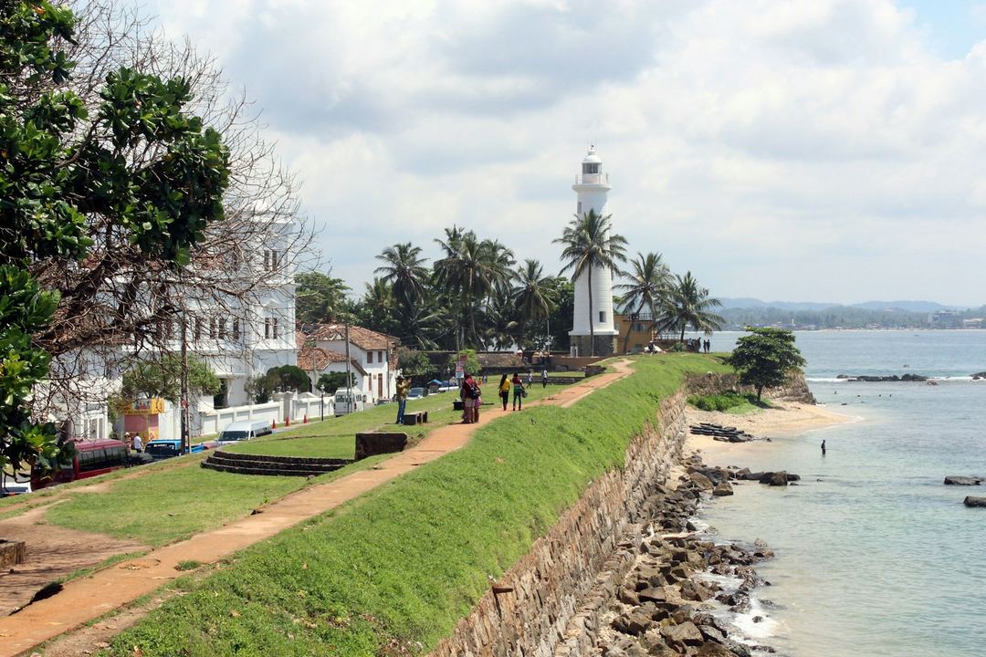 The white lighthouse on the ramparts of Galle Fort, with palms and the sea beside it.