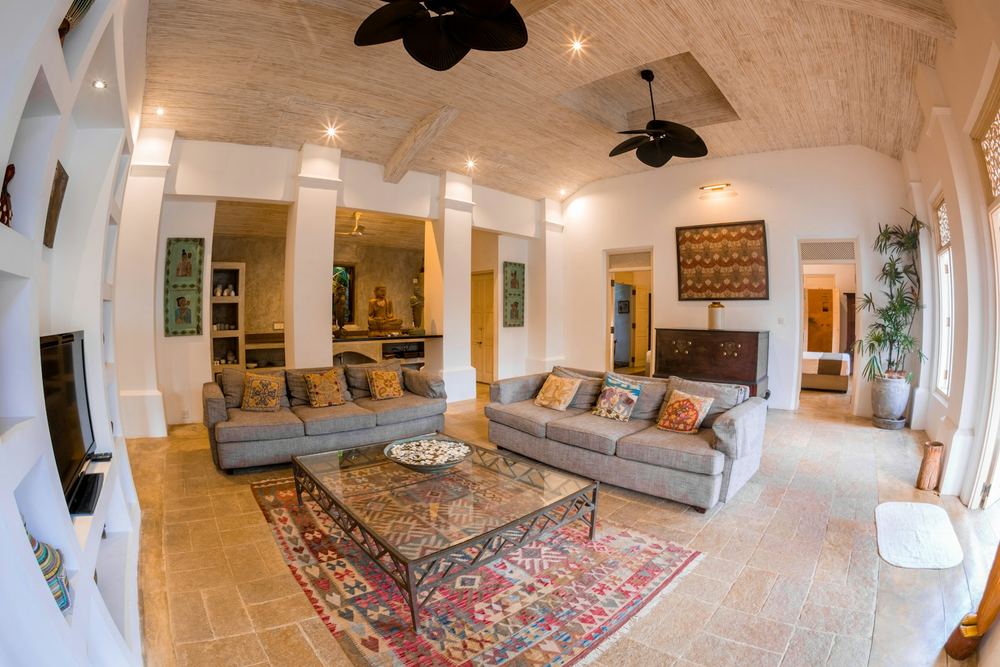 A high ceilinged lounge with patterned sofas, a carved chest and a ceiling fan.