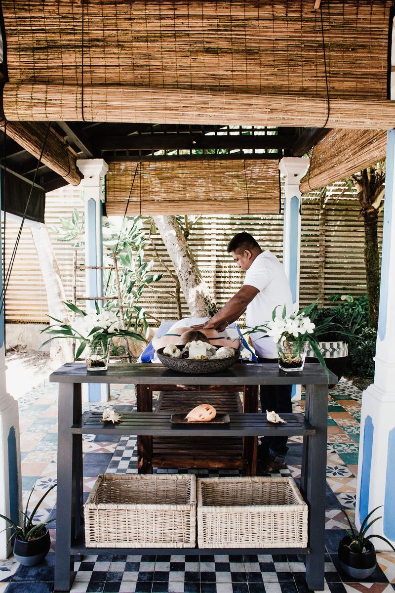 A therapist preparing oils at a table in an open pavilion, white pillars and a garden behind.