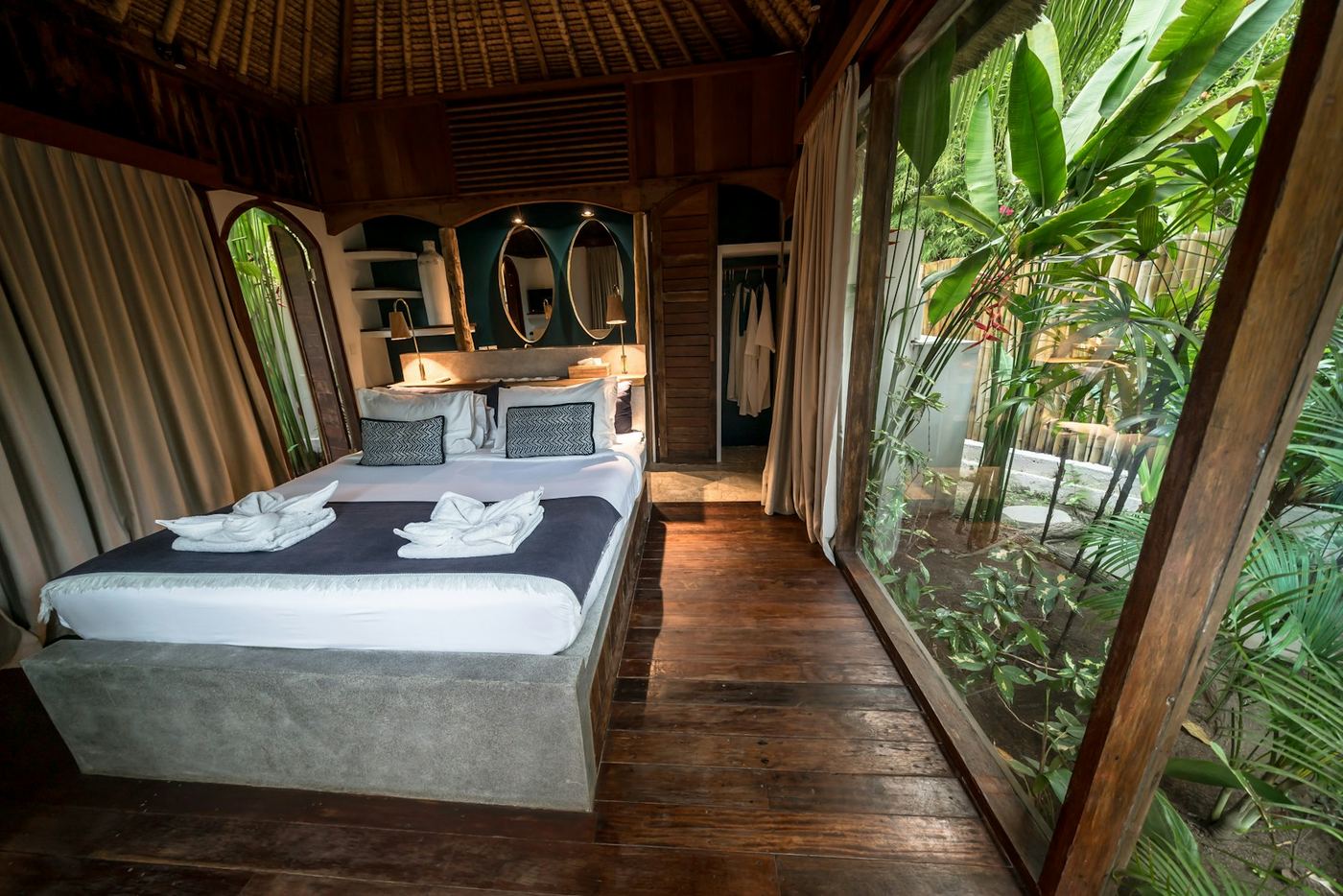 A bedroom with floor-to-ceiling glass on two sides, opening onto a dense tropical garden.