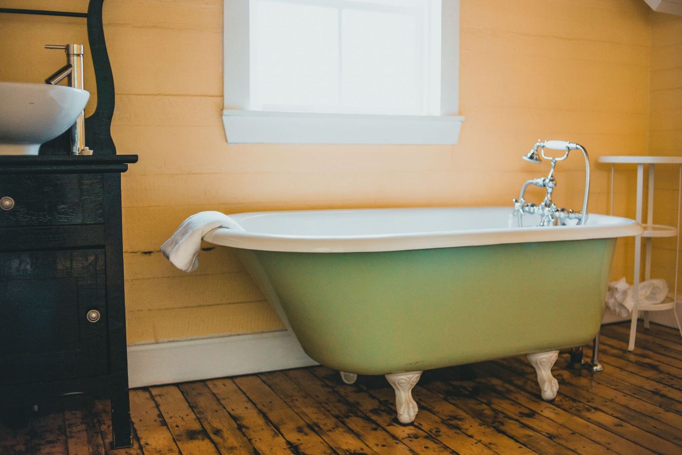 A green clawfoot bath on a timber floor, beneath a window.