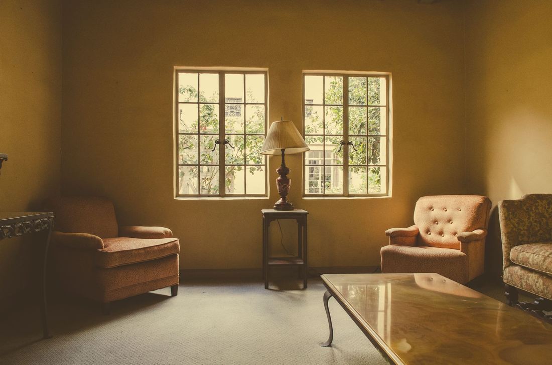 The sitting room in lamplight: two armchairs and a lamp between tall windows.