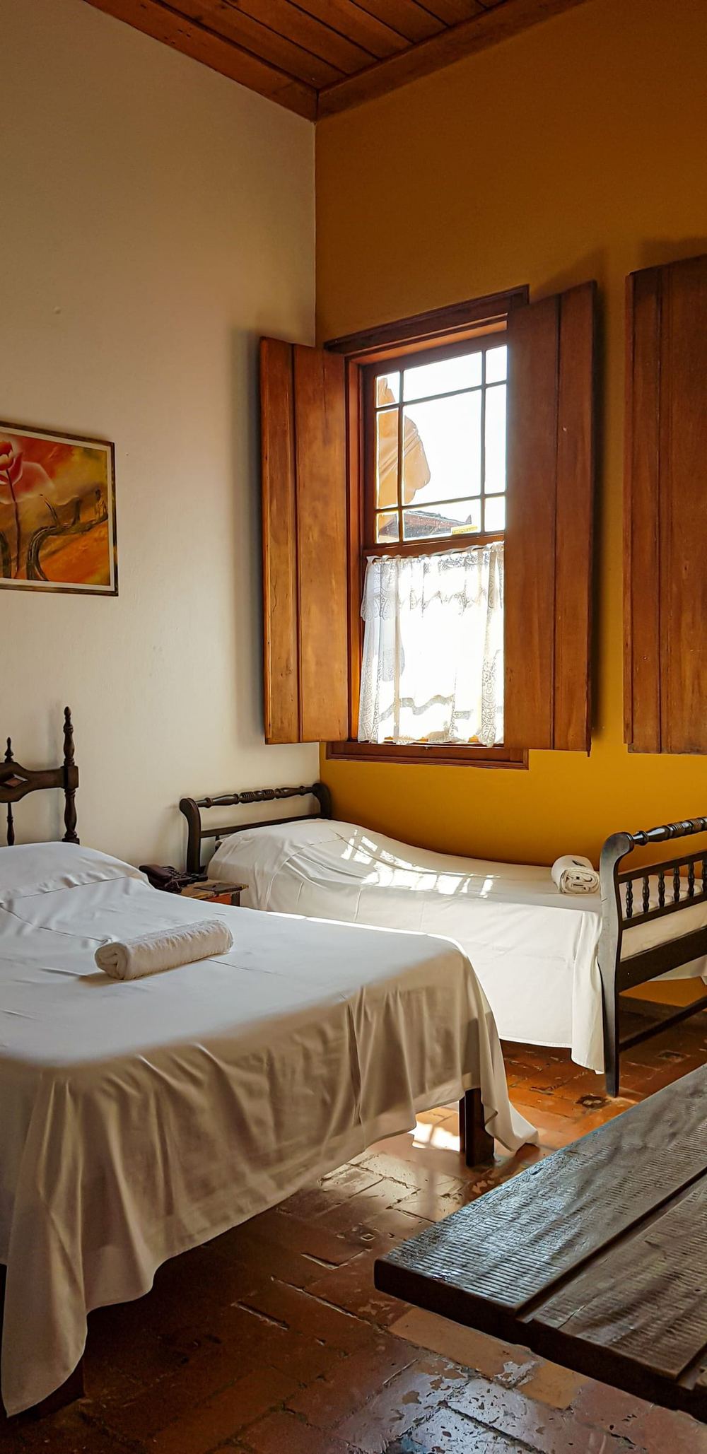 A bedroom with a yellow wall, twin beds and a shuttered window open to the light.