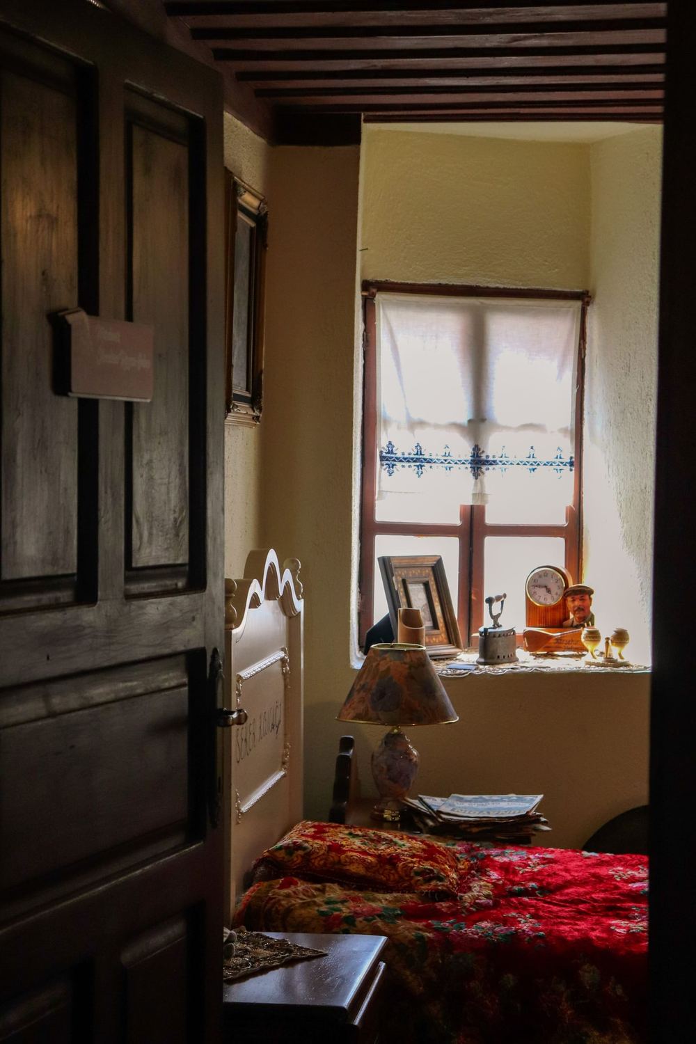 Through a heavy timber door: a room with a lamp by the window and a red bedspread.