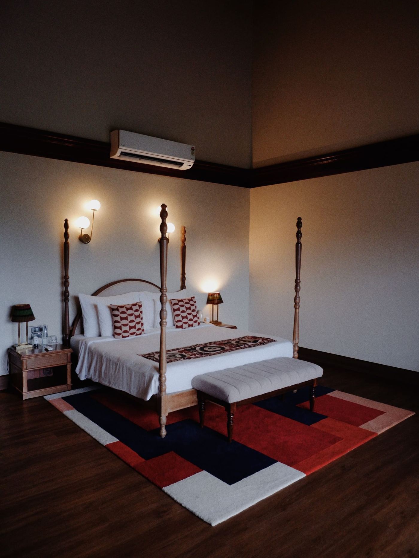 A four-poster bed lamplit in a high ceilinged room, a red rug at its foot.