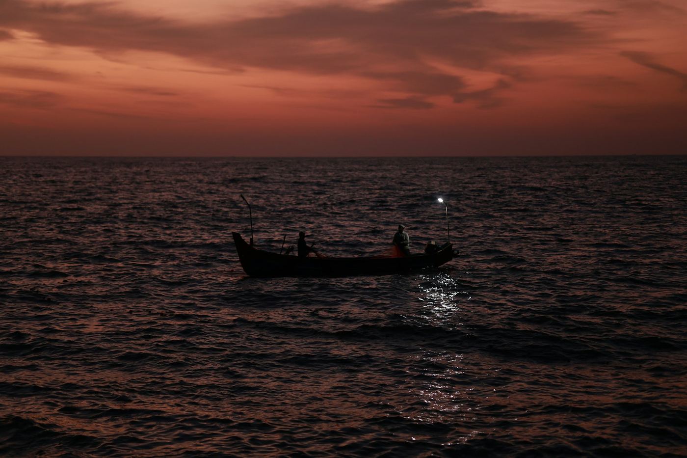 A small boat crossing dark water under a warm orange sky, two people aboard.