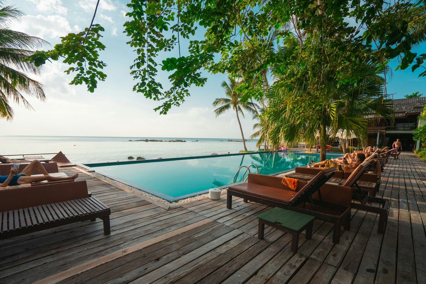 A timber deck with loungers beside an infinity pool, trees overhead and the sea beyond.