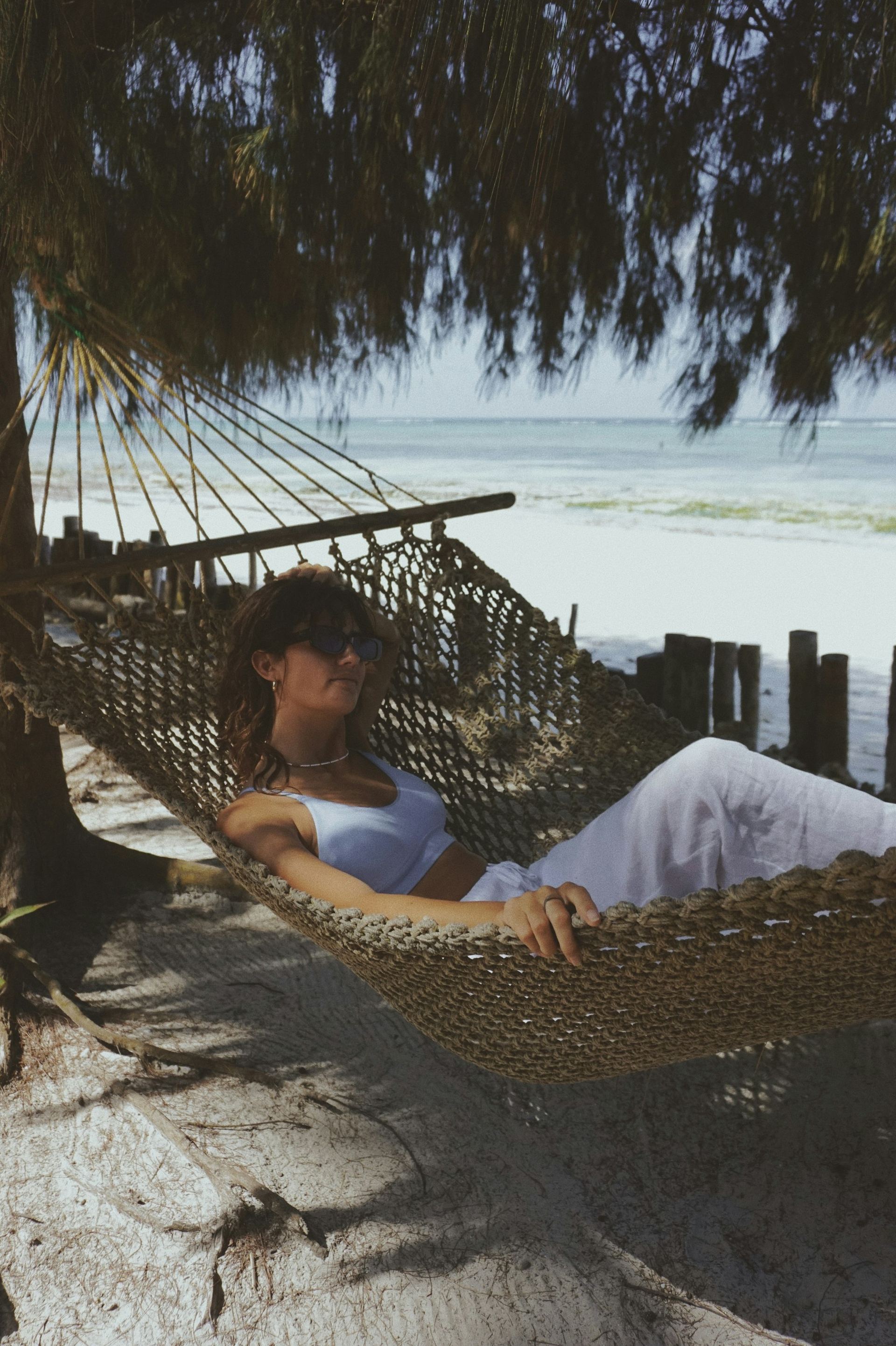A woman lying back in a rope hammock on a beach, eyes closed, the sea behind her.