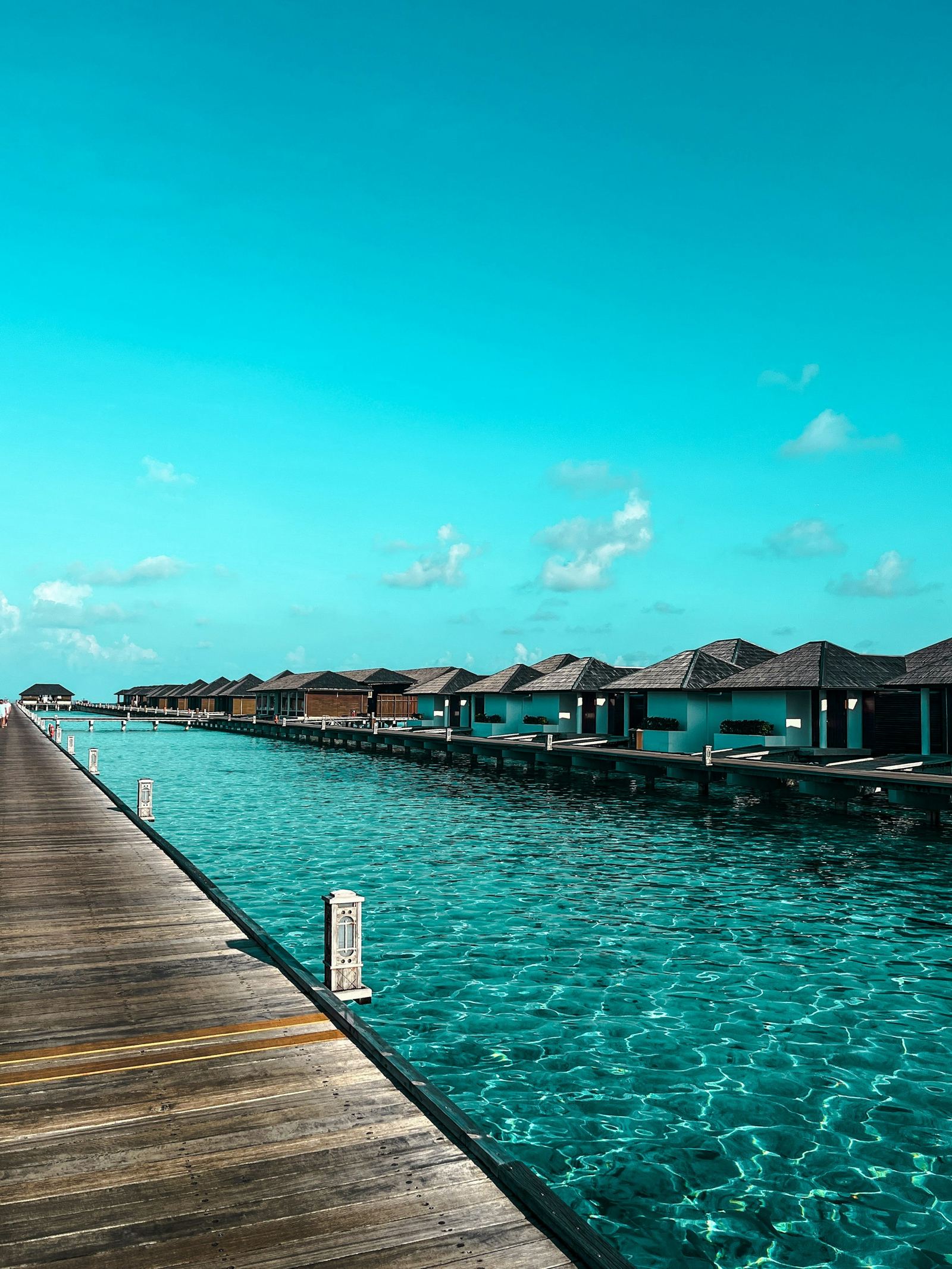 A timber boardwalk leading past a line of water villas over bright blue water.