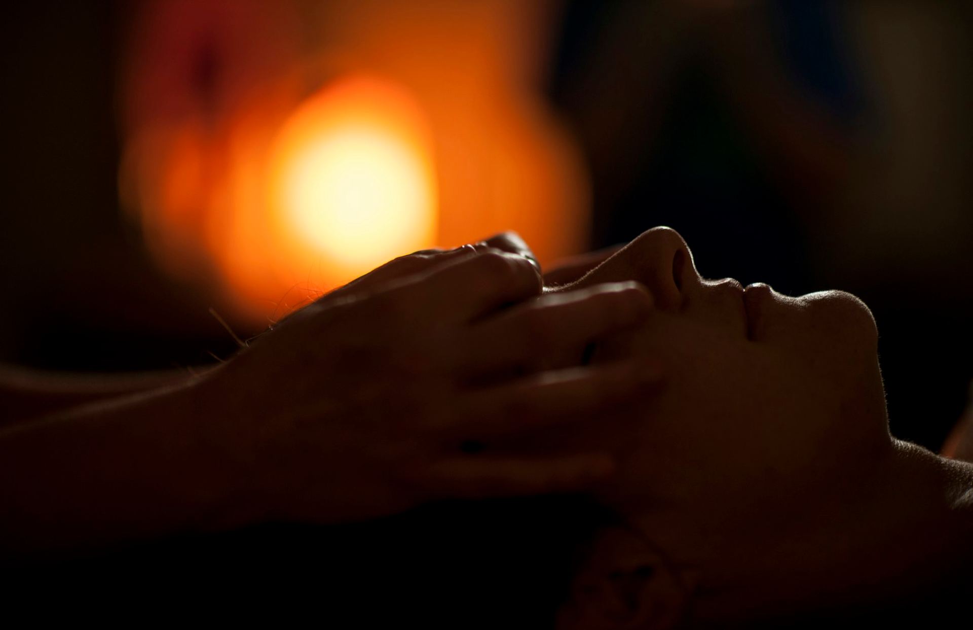 Hands resting on a guest’s face during a massage, a warm orange light glowing behind.