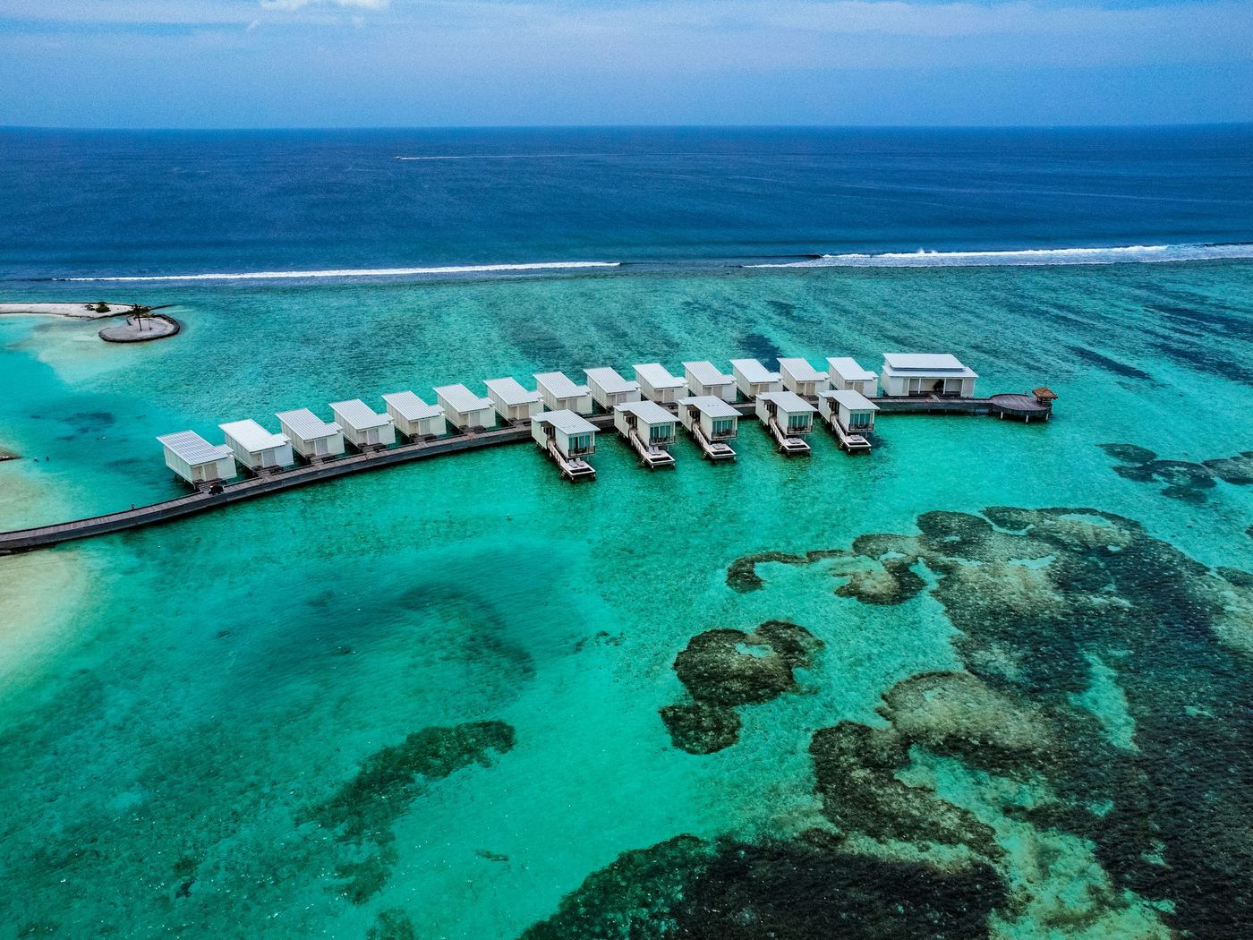 A row of villas on stilts reaching out over clear turquoise water and dark reef, from the air.