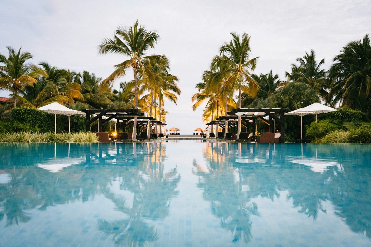 An infinity pool at a tropical hotel, palms and cabanas reflected in it, the sea beyond.