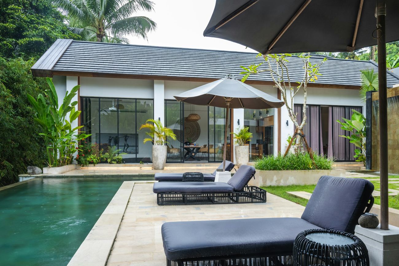 A white villa with a dark shingled roof and glass doors onto a long green pool, sun loungers under umbrellas on the terrace, in bright overcast light