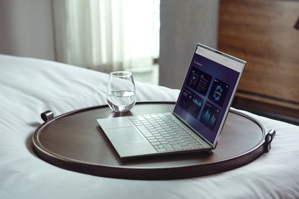 A laptop on a round wooden tray on a freshly made hotel bed.