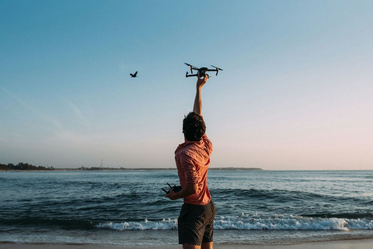 A man on the shore at dusk holding a drone up to launch it over the sea.
