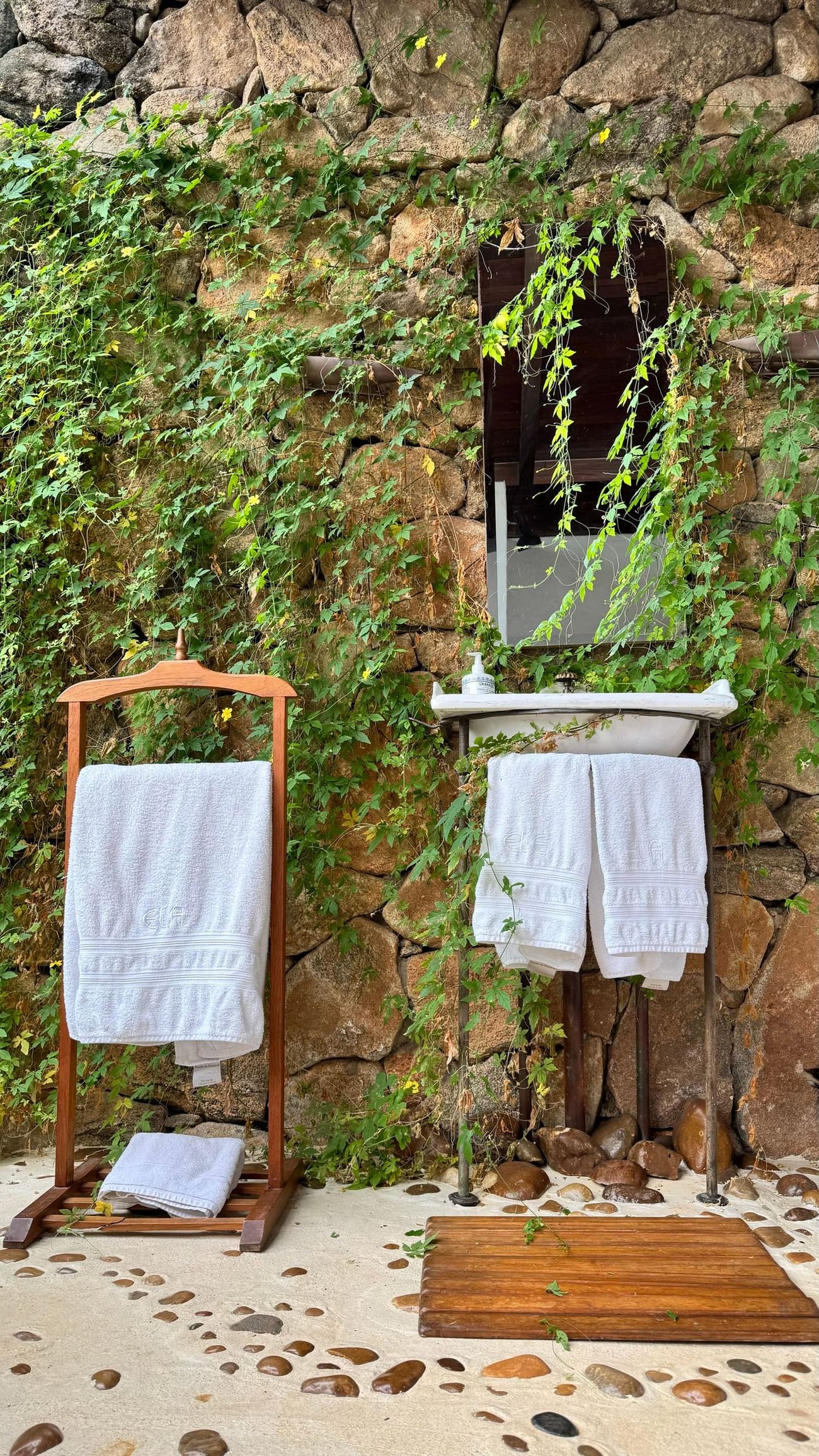An outdoor shower against a stone wall grown over with creeper, white towels on a timber rack.