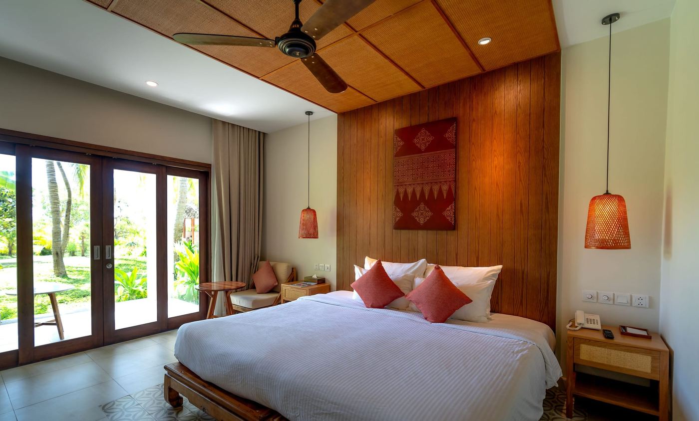 A bedroom with a timber wall behind the bed, a ceiling fan and glass doors onto green.